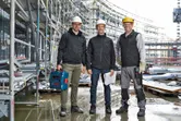安全装備を身に着けた 3 人の作業員が建設現場に立っており、1 人は電動工具を持っています。