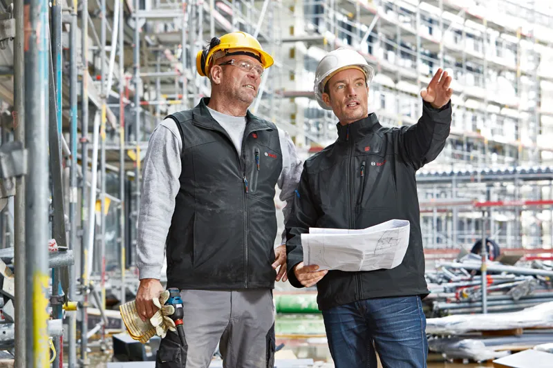 安全装備を身に着けた2人の作業員が建設現場で建築計画について話し合っている。.