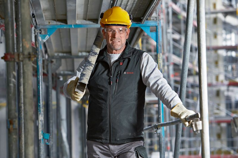 安全装備と加熱ベストを着用した人が建設現場で金属の梁を運んでいます。.