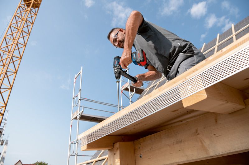 安全装備を身に着けた人が、建設中の建物の木製の梁にドリルで穴を開けています。.