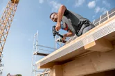 安全装備を身に着けた人が、建設中の建物の木製の梁にドリルで穴を開けています。.