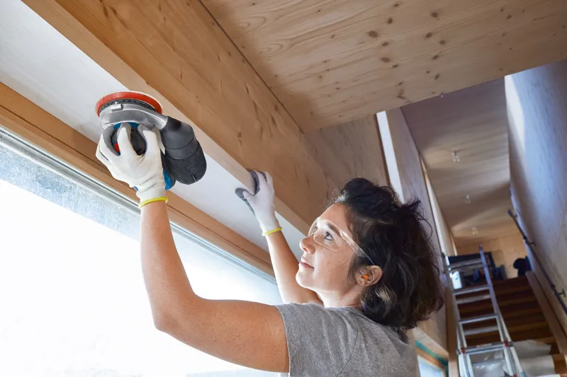 安全装備を身に着けた人がサンダーを使って屋内で木の梁を滑らかにしています。.