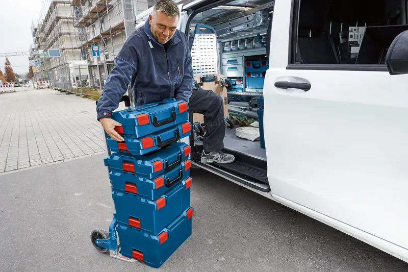安全装備を身に着けた人が、作業車の横にある台車に積み重ねた工具箱を積み込んでいます。