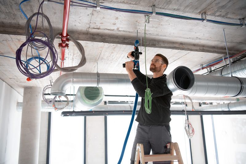 安全装置を装着した人が、はしごの上で FlexiClick アダプターを頭上にしてドリルを使用しています。