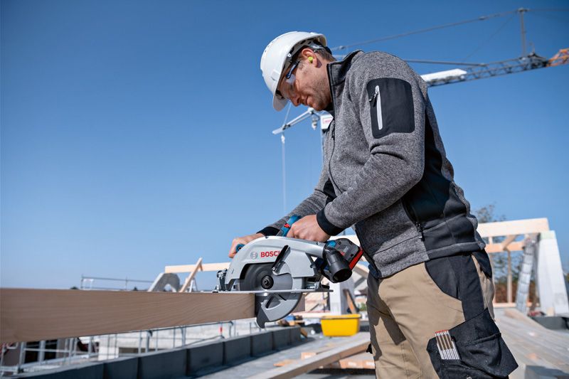 安全装備を身に着けた人が建設現場でコードレス丸鋸を使って木材を切っています。