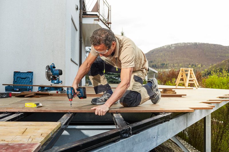 安全装備を身に着けた人が、屋外パティオのフレームにデッキ板をドリルで穴を開けています。.