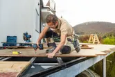 安全装備を身に着けた人が、屋外パティオのフレームにデッキ板をドリルで穴を開けています。.