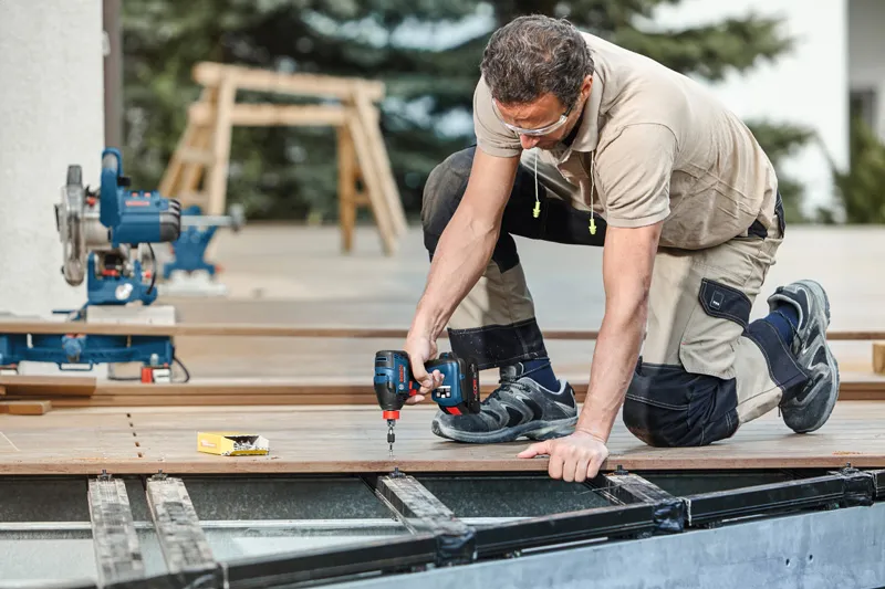 安全装備を身に着けた人が電動ドリルを使用して屋外のデッキ板を固定しています。.