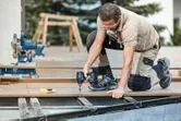 安全装備を身に着けた人が電動ドリルを使用して屋外のデッキ板を固定しています。