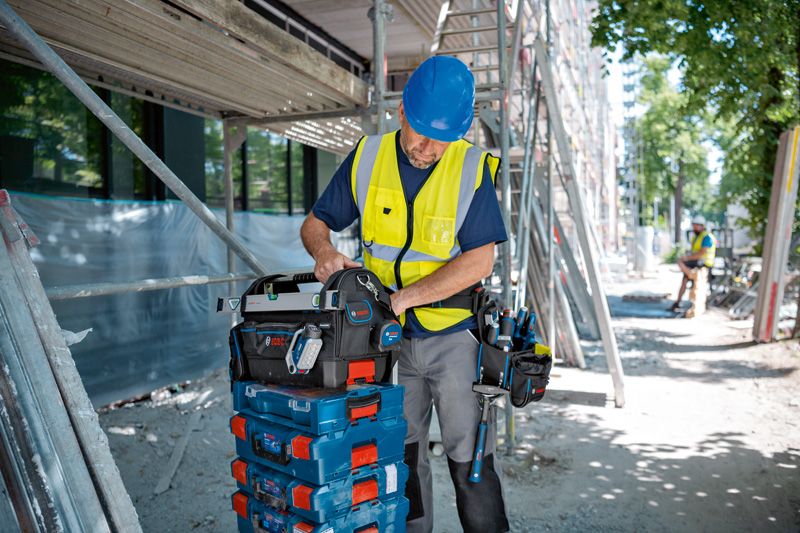建設現場で、安全装備を身に着けた人が積み重ねられた工具ケースの上に工具バッグを整理しています。.
