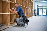 安全装備を身に着けた人が明るい部屋で木製の棚を組み立てています。