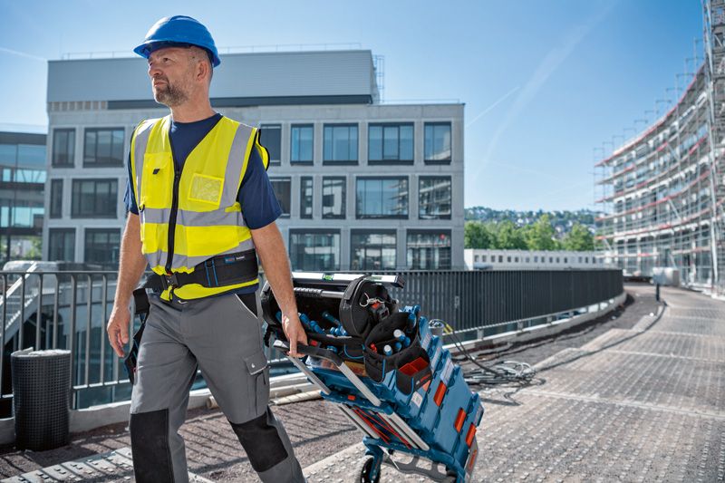 安全装備を身に着けた人が、建設現場で荷物を積んだ工具カートと工具バッグを引いています。.