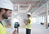 建設中の建物内で、安全装備を装着した2人がレーザー測定器とタブレットを使用しています。