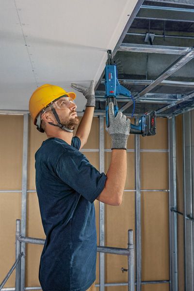 安全装備を身に着けた人が、電動工具を使って天井のフレームに石膏ボードを取り付けている。