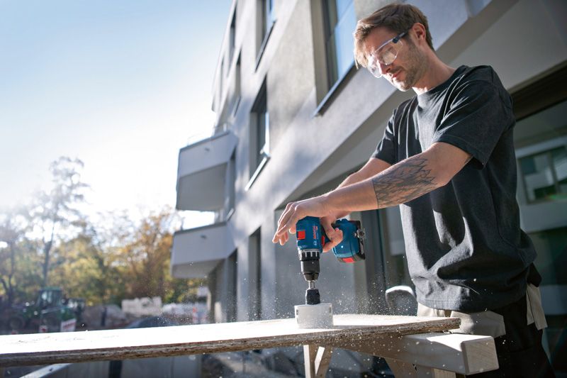安全装備を身に着けた人が屋外で電動ドリルを使って木材に穴を開けています。.