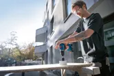 安全装備を身に着けた人が屋外で電動ドリルを使って木材に穴を開けています。.
