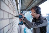 安全装置を装着した人がコードレス電動工具でレンガの壁に穴を開けています。