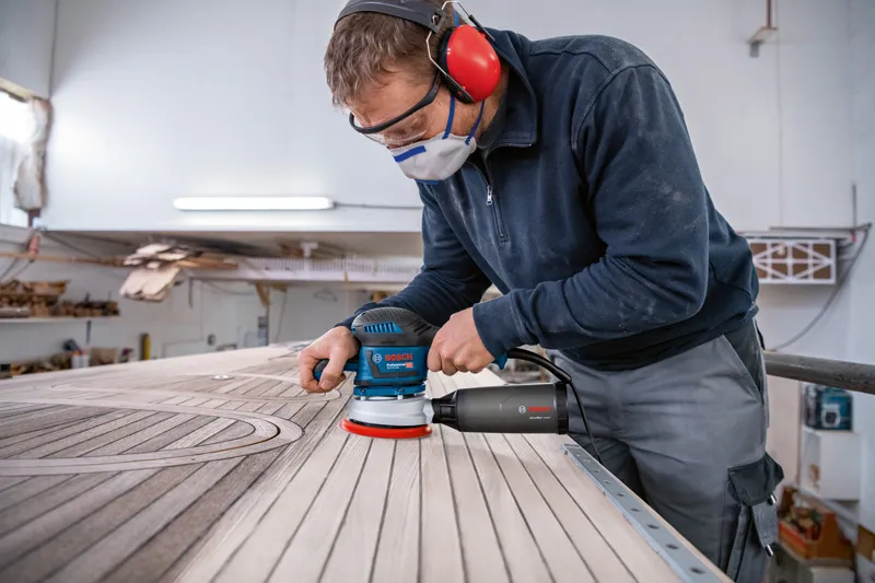 安全装備を身に着けた人がランダムオービットサンダーで木の板を研磨しています。.