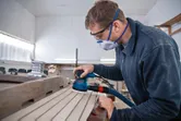 安全装備を身に着けた人がランダムオービットサンダーで木の板を研磨しています。