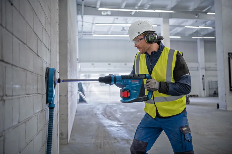 安全装置を装着した人が、コードレス回転ハンマーを使用してコンクリートの壁に穴を開けています。.