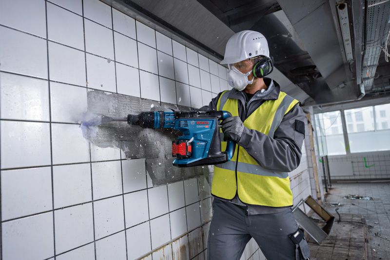 安全装備を身に着けた人が、コードレス回転ハンマーを使用して壁からタイルを取り外します。.