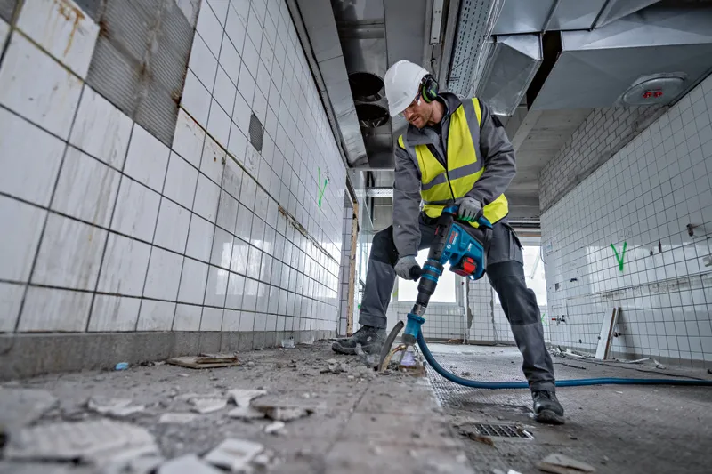 安全装備を身に着けた人がコードレス回転ハンマーでタイル張りの床を壊しています。.