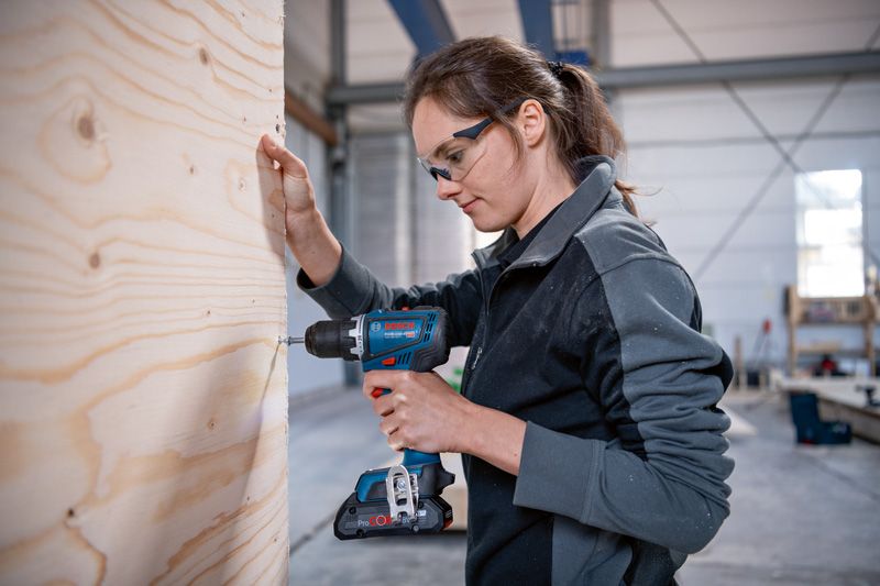 安全装備を身に着けた人が作業場で木の板に穴を開けています。.