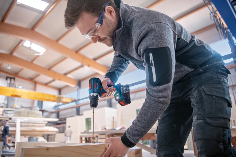 安全装置を装着した人がコードレス電動工具で木材に穴を開けています。.