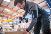 安全装置を装着した人がコードレス電動工具で木材に穴を開けています。.