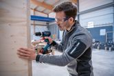 安全装備を身に着けた人が作業場内で木の板にドリルで穴を開けている。.