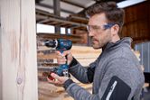 安全装備を身に着けた人が作業場で木の梁に穴を開けています。