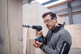 安全装置を装着した人がコードレスドリルドライバーを使用して木材に穴を開けています。.
