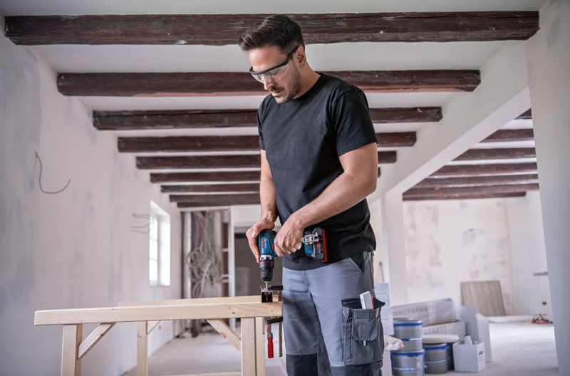 安全装備を身に着けた人が屋内建設現場で木材にドリルで穴を開けています。.
