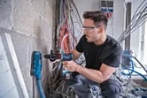 安全装備を身に着けた人が、集塵機を使ってコンクリートの壁に穴を開けています。.