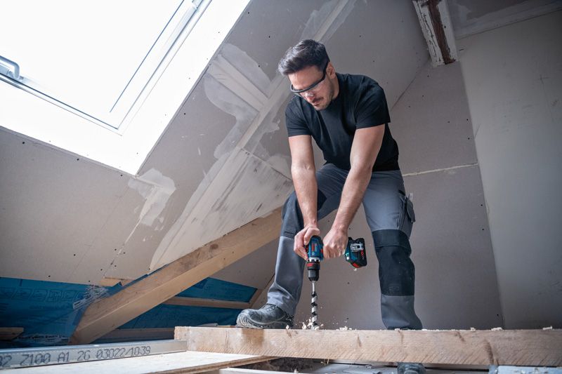 安全装備を身に着けた人が、完成途中の屋根裏部屋の木の梁にドリルで穴を開けている。.