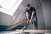 安全装備を身に着けた人が、完成途中の屋根裏部屋の木の梁にドリルで穴を開けている。.