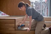 安全装備を身に着けた人がコードレスオービタルサンダーで木製の階段を研磨しています。.