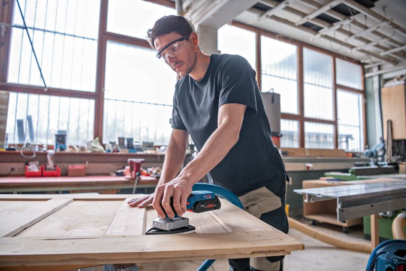 安全装備を身に着けた人がコードレスオービタルサンダーで木製のドアを研磨しています。.