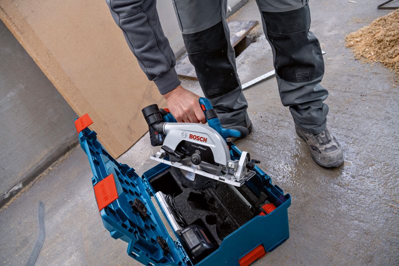 安全装備を身に着けた人が作業場の床にある工具キットから丸鋸を持ち上げています。.