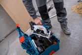 安全装備を身に着けた人が作業場の床にある工具キットから丸鋸を持ち上げています。.