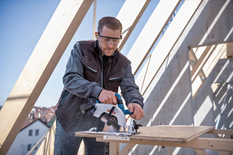 安全装置を装着した人が建設現場で丸鋸を使用して合板を切断しています。.