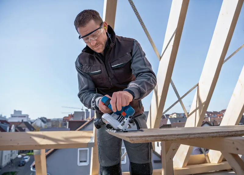 安全装備を身に着けた人が建設現場でコードレス丸鋸を使って木材を切っています。.