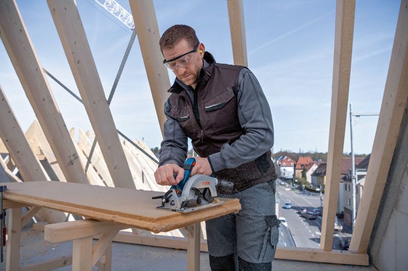 建設現場で安全装備を身に着けた人が丸鋸で木の板を切っている。.