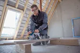 安全装備を身に着けた人が、未完成の屋根裏部屋で丸鋸を使って木の板を切っています。