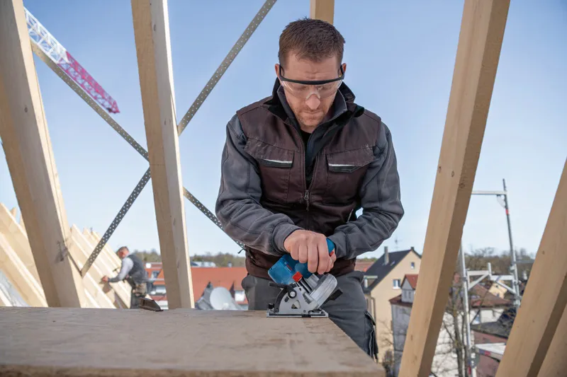 安全装備を身に着けた人が建設現場で電動のこぎりを使って木材を切断しています。.