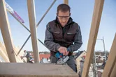 安全装備を身に着けた人が建設現場で電動のこぎりを使って木材を切断しています。.