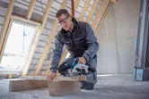 安全装備を身に着けた人が屋根裏で丸鋸を使って木の梁を切っています。.