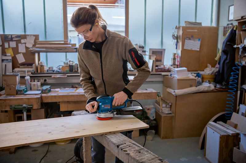 安全装置を装着した人がランダムオービットサンダーで木の板を研磨している。.