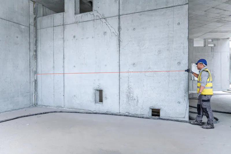 安全装備をつけた人がレーザー測定器でコンクリートの壁を測定しています。.
