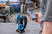 作業場で、スマートフォンを使ってコードレスジグソーを調整している人。